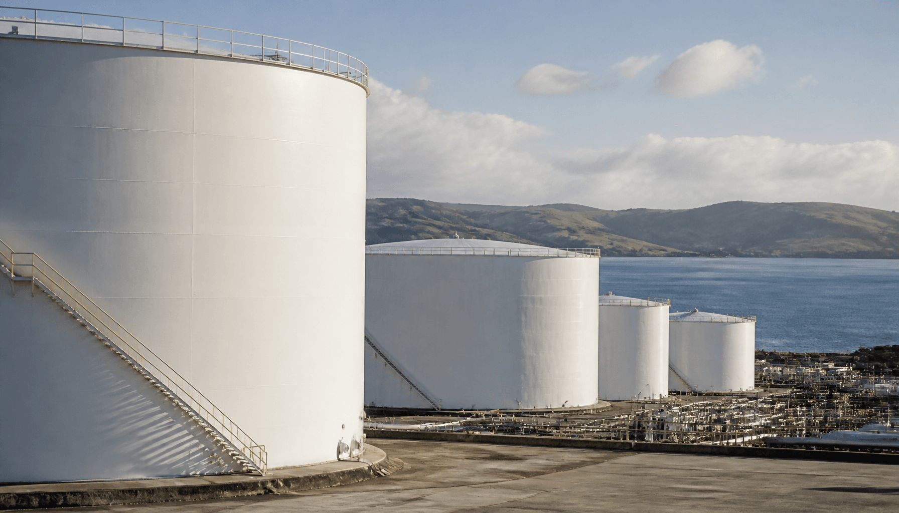 Large white diesel storage tanks at Marsden Point Energy Precinct, Northland, with calm harbour water and rolling hills behind
